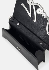 Pochette en cuir noir texturé avec rabat ouvert montrant un intérieur spacieux et des lettres décoratives en métal argenté à l'intérieur du rabat.
