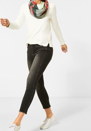 Kobieta ubrana w czarne obcisłe jeansy z ćwiekami, białą bluzkę z długim rękawem, kwiecisty szal i białe trampki, idąca na białym tle.