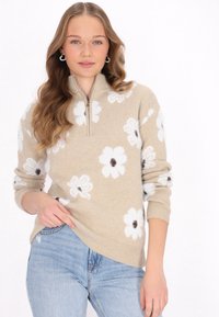Pulover bej cu fermoar pe un sfert, decorat cu modele florale albe și centre întunecate. Mânecile și tivul riglate, purtat cu blugi deschiși.