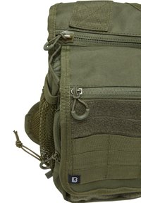 Olivgrön taktikpåse tillverkad av slitstarkt tyg, med flera zip-pockets, nätinslag och remmar för fästen.