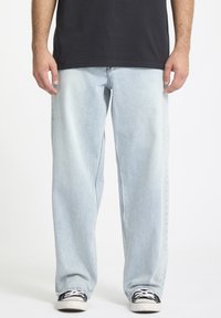 Mann i lyseblå løse jeans, svarte sneakers med hvite snørebånd, og en svart t-skjorte mot en hvit bakgrunn.