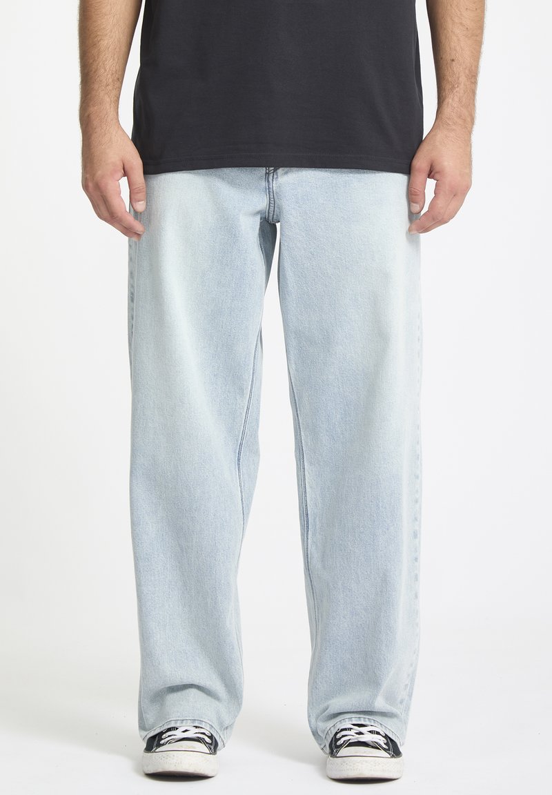 Mann i lyseblå løse jeans, svarte sneakers med hvite snørebånd, og en svart t-skjorte mot en hvit bakgrunn.