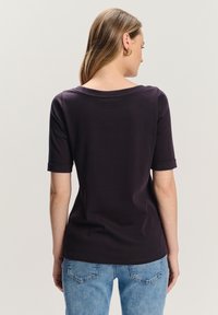 Kobieta z długimi włosami, ubrana w ciemnofioletową koszulę z krótkim rękawem i jasnoniebieskie dżinsy, stojąca tyłem na tle jasnego, jednolitego tła.