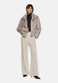 Faux-Fur-Jacke in Hellgrau mit hohem Kragen, Reißverschluss und oversized Schnitt, kombiniert mit cremefarbenen weit geschnittenen Hosen und schwarzen High Heels.