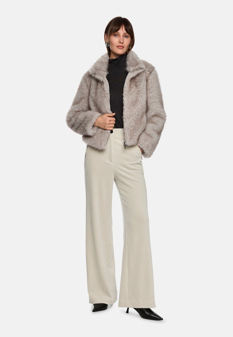 Faux-Fur-Jacke in Hellgrau mit hohem Kragen, Reißverschluss und oversized Schnitt, kombiniert mit cremefarbenen weit geschnittenen Hosen und schwarzen High Heels.