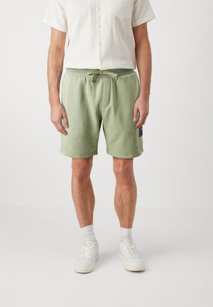 Shorts en coton vert avec une taille élastique et un cordon de serrage, dotés de poches latérales et d'une étiquette de marque sur la jambe gauche.