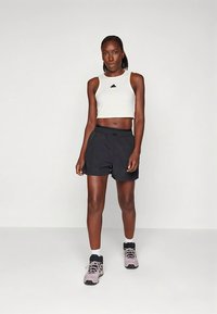 Sieviete stāv, tērpusies baltā bezdelņu kreklā, melnās šortos, baltās zeķēs un pelēkos sporta apavos uz vienkārša gaiša fona.