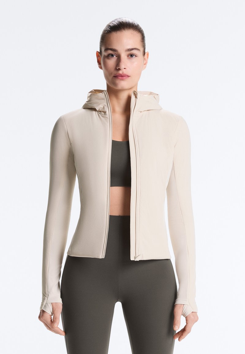 Femme aux cheveux tirés en arrière portant une veste à capuche beige zippée par-dessus un soutien-gorge de sport sombre et un legging, debout devant un fond blanc.