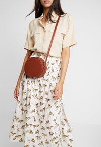 Borsa a tracolla in pelle marrone chiaro con forma rotonda e zip frontale, indossata sopra una gonna bianca con una stampa di cavalli ripetuta in marrone.