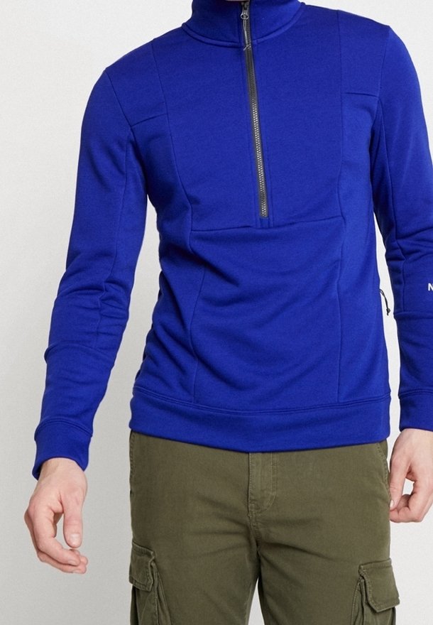Homme portant un pull à manches longues bleu vif avec une fermeture éclair partielle et un pantalon cargo vert olive, debout les bras légèrement écartés.