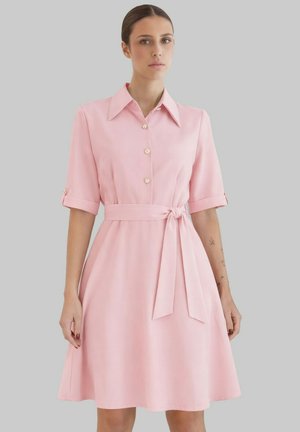 Femme portant une robe rose mi-longue avec manches courtes, devant boutonné, col large et ceinture à la taille, sur un fond gris uni.