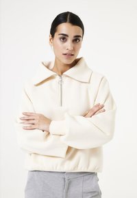 Sudadera color crema con un cuello alto, cremallera de medio recorrido en la parte delantera y puños elásticos. Tejido suave y texturizado con un corte relajado.