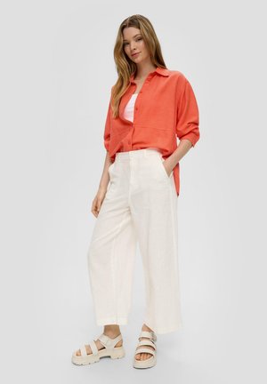 Ein korallenfarbener Leinen-Button-Up-Hemd über einem weißen Tanktop, kombiniert mit weit geschnittenen creamfarbenen Hose und beigen, klobigen Sandalen mit verstellbarem Riemen.