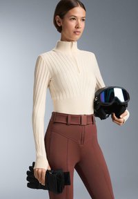 Krém színű bordázott, zippzáros pulóver sötétbarna, testhezálló nadrággal párosítva. Az egyik kezében fekete sígömb és kesztyű van.
