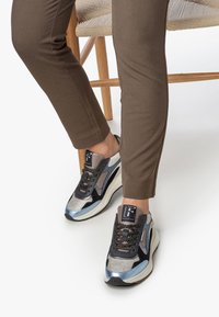 Vaatteet ruskeissa housuissa istuu punospuu tuolilla, jalassa mustat, hopeiset, siniset ja beige-kuosiset tennarit, joissa on paksut valkoiset pohjat.