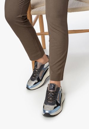 Beine in braunen Hosen, die auf einem geflochtenen Holzstuhl sitzen, mit schwarzen, silbernen, blauen und beigen gemusterten Sneakers mit dicken weißen Sohlen.