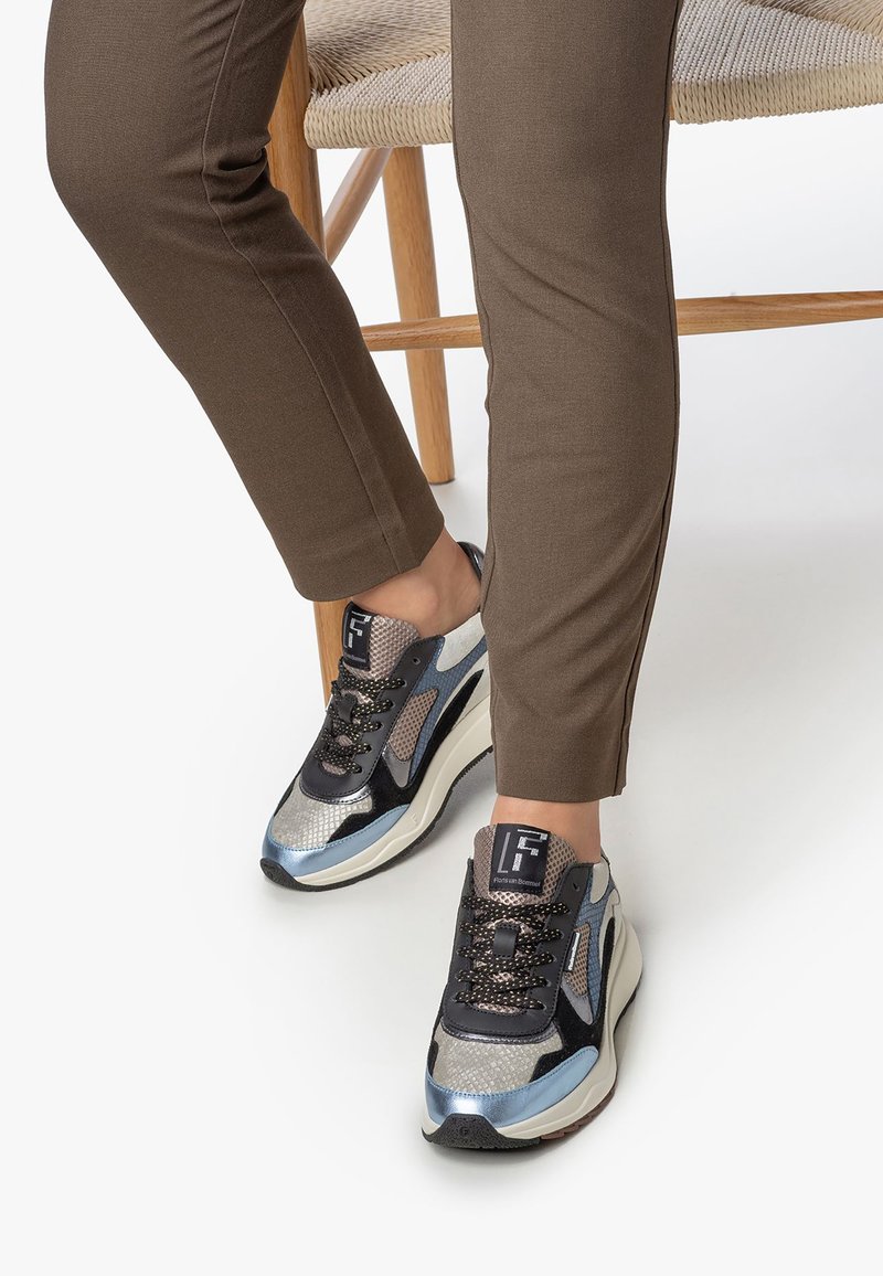 Vaatteet ruskeissa housuissa istuu punospuu tuolilla, jalassa mustat, hopeiset, siniset ja beige-kuosiset tennarit, joissa on paksut valkoiset pohjat.