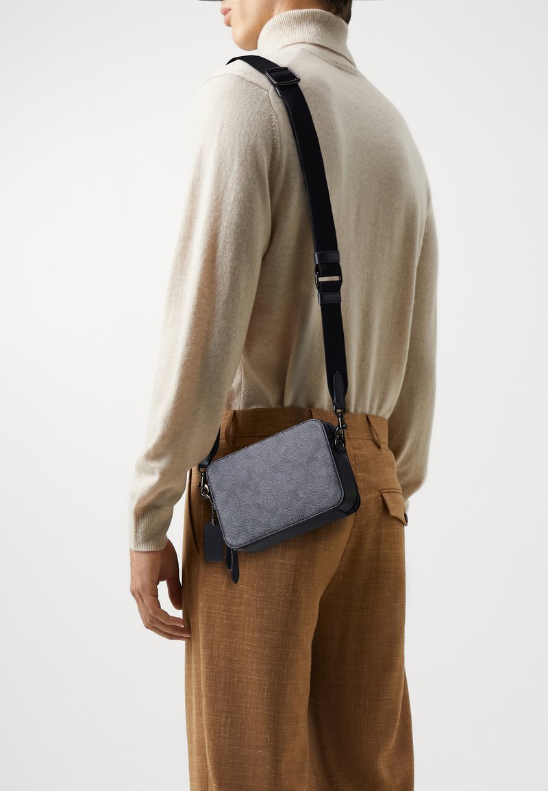 Bolso cruzado gris con superficie texturizada, detalles en cuero negro y correa ajustable, llevado sobre un suéter beige y pantalones marrones.