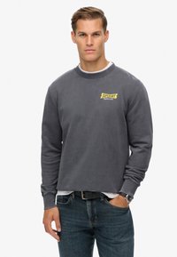 Hombre vistiendo un sudadera Superdry gris oscuro, jeans azules, cinturón negro y reloj de pulsera, de pie con una mano en el bolsillo contra un fondo blanco.