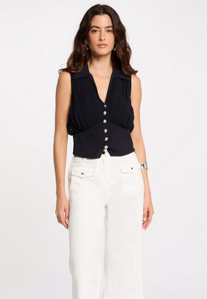 Mujer con una blusa negra sin mangas con botones y pantalones blancos de talle alto con bolsillos delanteros, de pie frente a un fondo sencillo.