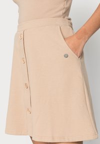 Saia skater bege feita de algodão macio, com cintura, fecho de botão frontal, bolsos laterais e um pequeno detalhe metálico perto do bolso.