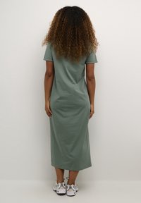 Olivnozielona, do kolan, dopasowana sukienka T-shirt z krótkimi rękawami, wykonana z gładkiego materiału, w połączeniu z białymi i szarymi butami sportowymi.