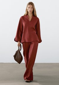 Haut en lin cramoisi avec un col en V et des manches larges, associé à un pantalon large assorti. Le mannequin porte un sac marron texturé.