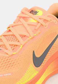 Orange löparsko med mesh-övre, svart Nike-swoosh, gula accenter, platta snörningar och mönstrad sula med synliga spår.