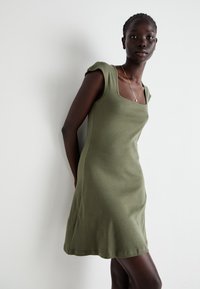 Vestuário verde oliva, ajustado, com decote quadrado e ombros curtos. Feito de um tecido macio com uma textura suave e bainha até ao joelho.