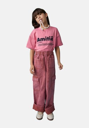 Enfant portant un t-shirt rose « Amitié » et un pantalon rose large, tenant un collier de bonbons enroulé autour de sa bouche sur un fond uni.