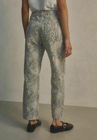 Slangenprint jeans in tinten grijs en groen, met een hoge taille, rechte pijpen en klassieke achterzakken, gecombineerd met zwarte platte schoenen.