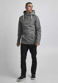Sudadera gris con cuello de bufanda, cordones blancos, bolsillo frontal y dobladillo acanalado. Combinada con pantalones negros y zapatillas oscuras.