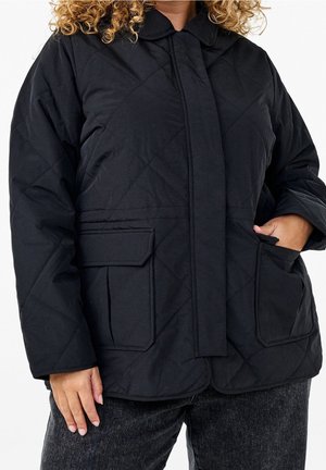 Personne portant une veste matelassée noire avec des poches à rabat, un col, et une main dans la poche, associée à un jean en denim foncé.