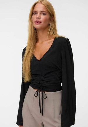 Femme aux longs cheveux blonds portant un haut noir froncé à col en V et un pantalon taille haute beige clair sur un fond blanc uni.