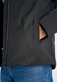 Svart ytterplagg med en slät yta, utrustad med blixtlås och en sidoficka. Kombinerat med blå denimjeans.