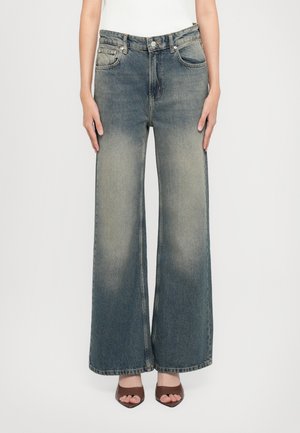 Woman wearing high-waist wide-leg faded blue jeans and brown heeled sandals, standing against a plain white background.