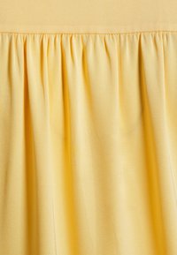 Tessuto giallo con una consistenza liscia, caratterizzato da pieghe verticali lungo il bordo superiore, creando un aspetto fluido in tutto il capo.
