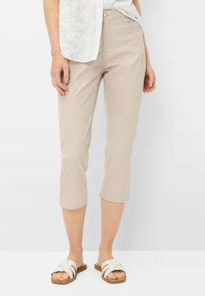 Vrouw die beige korte broek, witte kanten top en witte slippers draagt, staand tegen een witte achtergrond.