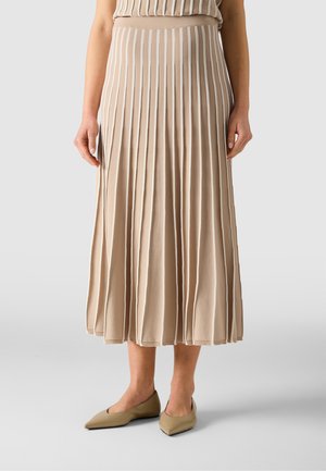 Kvinde iført en beige plisseret midi-nederdel kombineret med spidse beige flade sko, stående foran en ensartet lys grå baggrund.