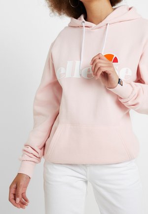 Persoon die een lichtroze hoodie draagt met witte trekkoorden en wit tekstlogo, gecombineerd met een witte broek, getoond vanaf de midden van het bovenlichaam naar beneden.
