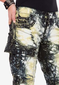 Pantalones de tie-dye en negro y amarillo con acentos de tachuelas y un acabado texturizado. Presentan un ajuste de tiro bajo y bolsillos laterales tipo cargo.