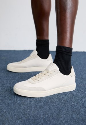 Kreminės odinės sportbačiai su tekstūruotu paviršiumi, apvaliu pirštu, paminkštintu apvadu ir lygiais raišteliais. Derinami su juodomis raukšlėtomis kojinėmis, padėtos ant mėlyno kilimo.