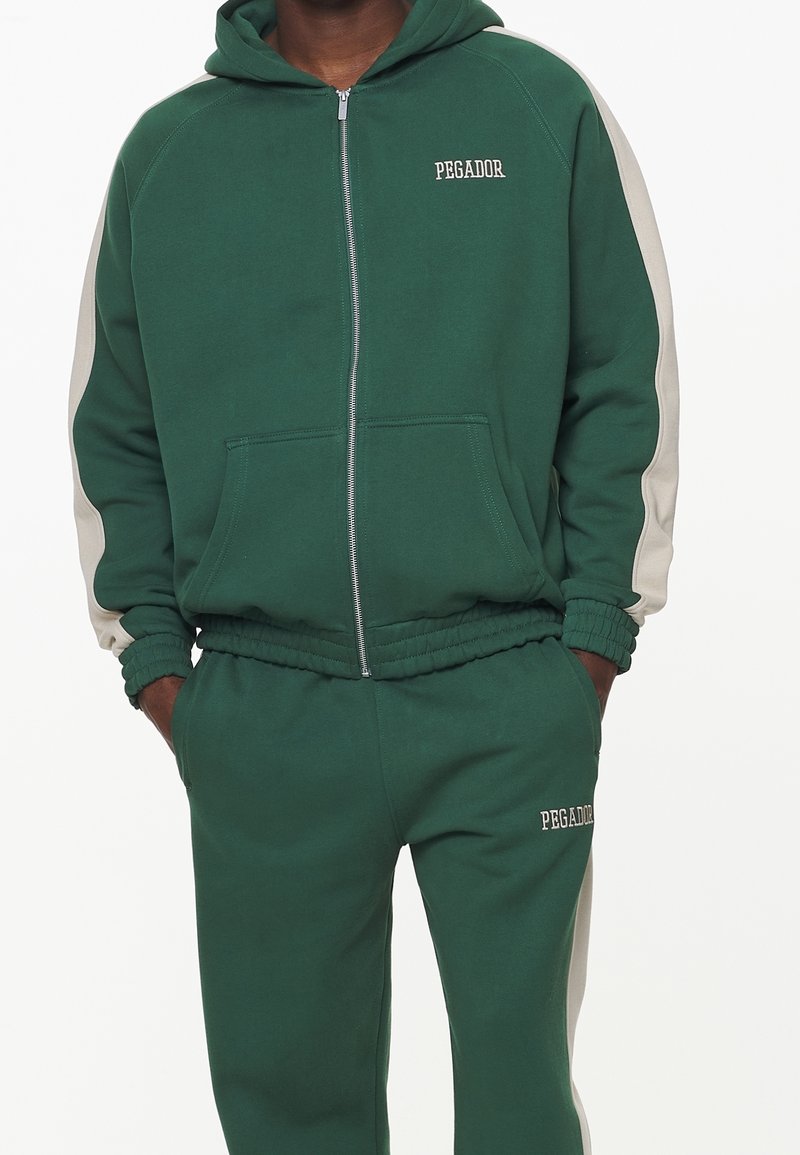 Felpa con zip verde con accenti laterali grigi e polsini a coste. Pantaloni verdi abbinati con vita elastica e ricamo del logo sulla gamba.