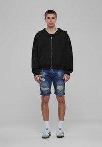 Čierna mikina na zips, potrhané modré džínsy s rozstrapkanými okrajmi, biele ponožky a šedé tenisky s čiernymi akcentmi. Jednoduchý, neformálny outfit.