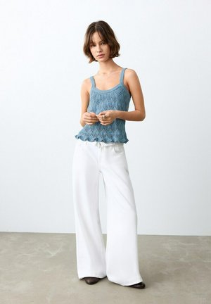 Femme aux cheveux courts bruns portant un débardeur bleu tricoté et un pantalon blanc à jambes larges, debout sur un sol gris devant un mur blanc.