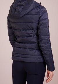 Giubbotto imbottito blu navy con cappuccio, con dettagli trapuntati e polsini elastici. Texture liscia e lucida con cerniere laterali.