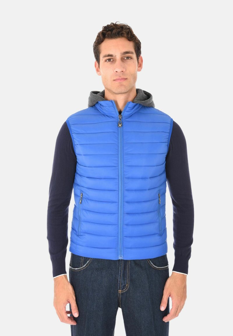 Uomo che indossa un gilet imbottito blu senza maniche su una maglietta nera a maniche lunghe e jeans scuri, in piedi davanti a uno sfondo bianco semplice.
