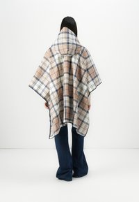 Person wearing a beige, white, and navy plaid hooded poncho with dark blue flared jeans, standing against a plain white background.
