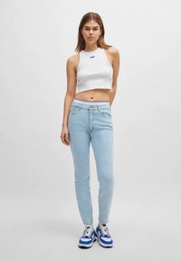 Jovem mulher vestindo uma blusa curta branca, jeans de cintura alta azul claro e tênis brancos e azuis, de pé contra um fundo simples.