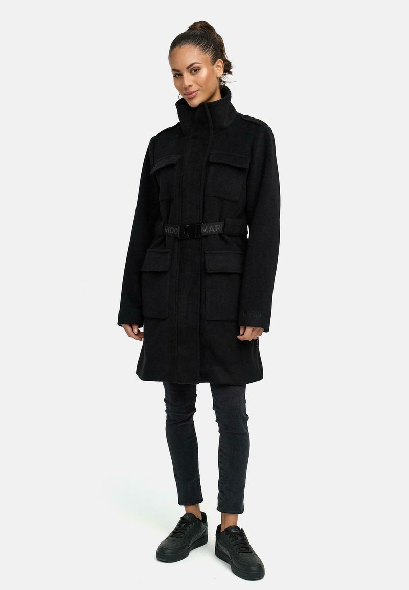 Cappotto in lana nera con colletto alto, più tasche frontali e una cintura. Abbinato a pantaloni slim fit scuri e sneaker nere.